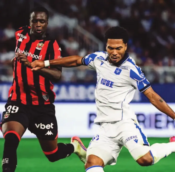 Un joueur rennais en maillot rouge et noir à la lutte avec un joueur d’Auxerre en blanc.